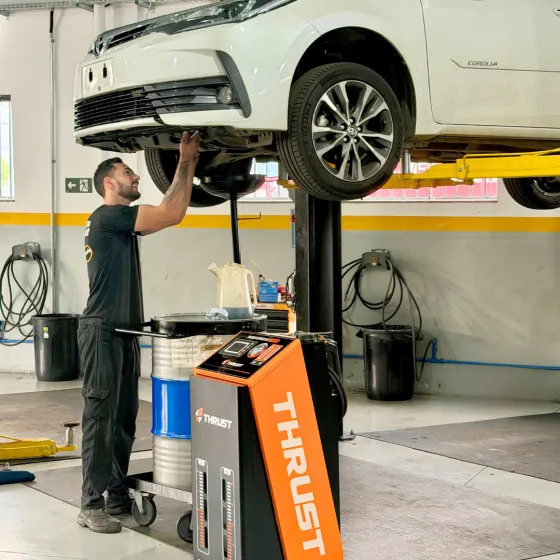 Toyota Corolla branco no elevador enquanto o técnico da Pneuscarmg trabalha sob o carro, ao lado do trocador de fluido ATF Thrust